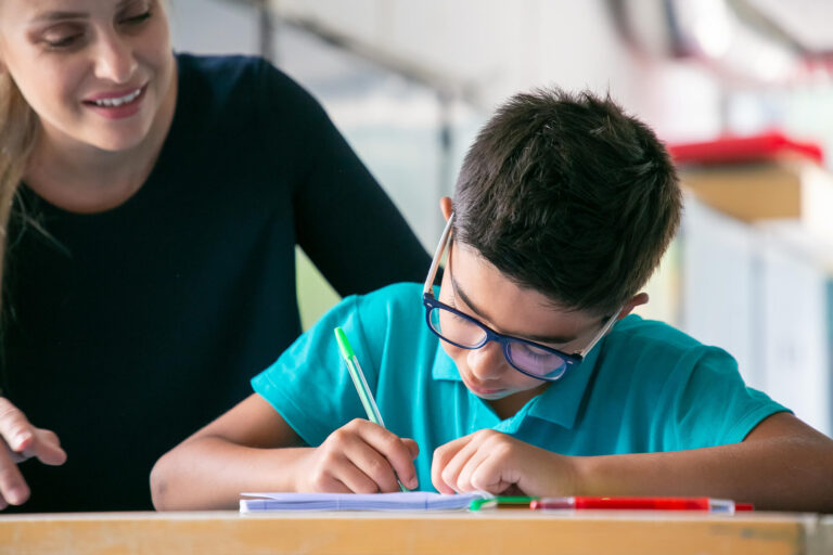 happy-school-teacher-watching-schoolboy-glasses-doing-task-class (1) (1)